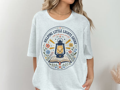 Light blue t-shirt with a circular graphic of a lantern over an open book and flowers.