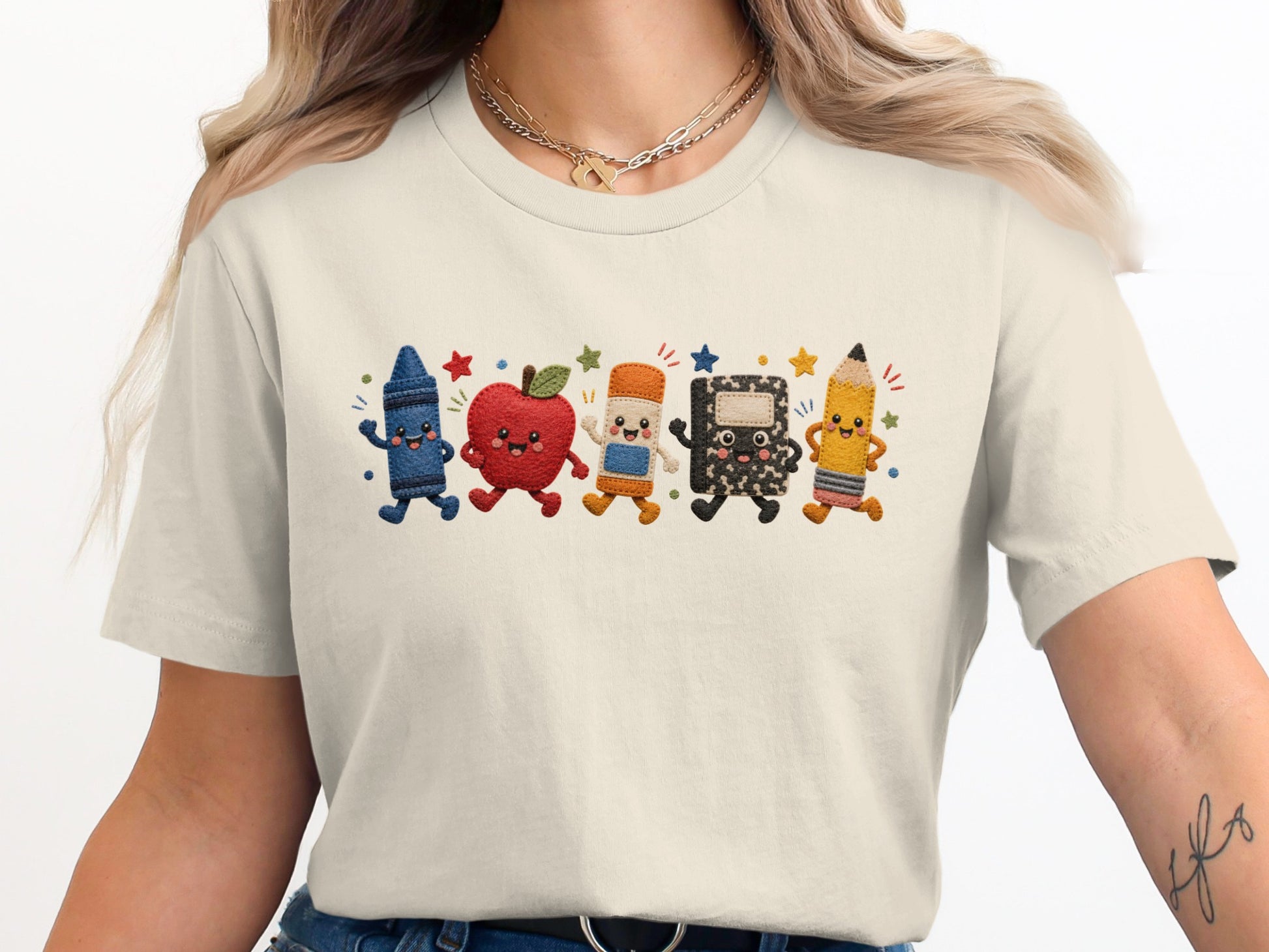 A cartoon crayon, apple, eraser, notebook, and pencil march across a cream shirt.