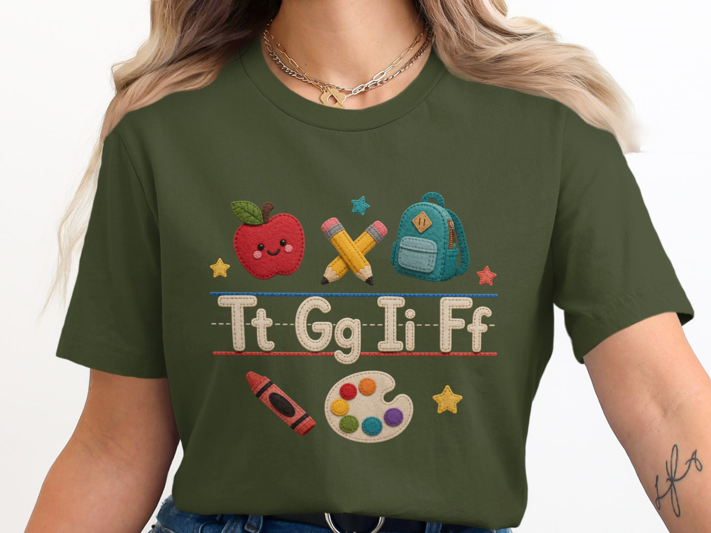 Green t-shirt with embroidered apple, pencils, backpack, crayon, paint palette, and letters Tt Gg Ii Ff.