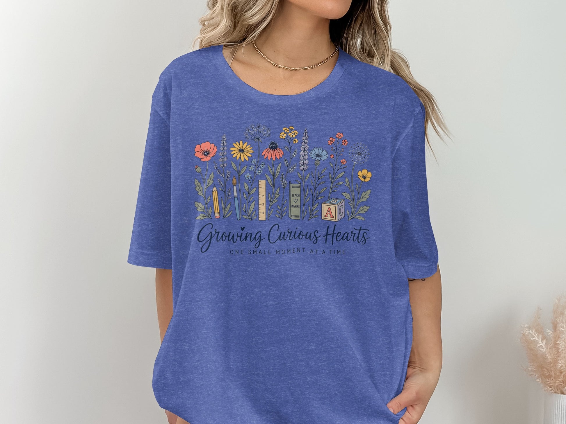 Blue t-shirt with floral design, pencil, ruler, book, and ABC blocks. Text reads "Growing Curious Hearts."