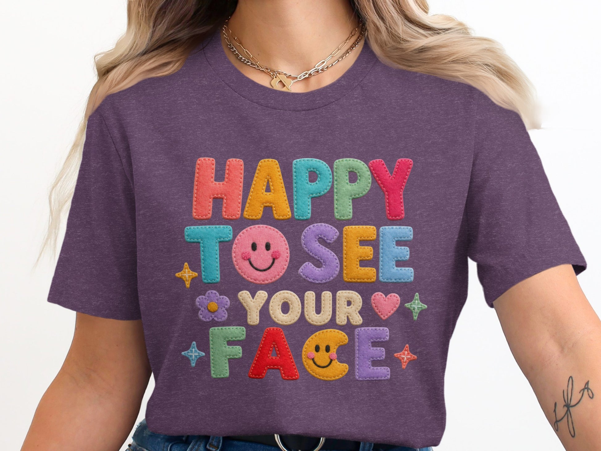 Purple t-shirt with "HAPPY TO SEE YOUR FACE" text in colorful embroidered letters.