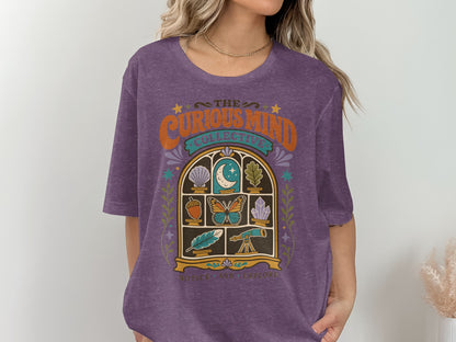 Purple t-shirt with "The Curious Mind Collective" and window illustration.