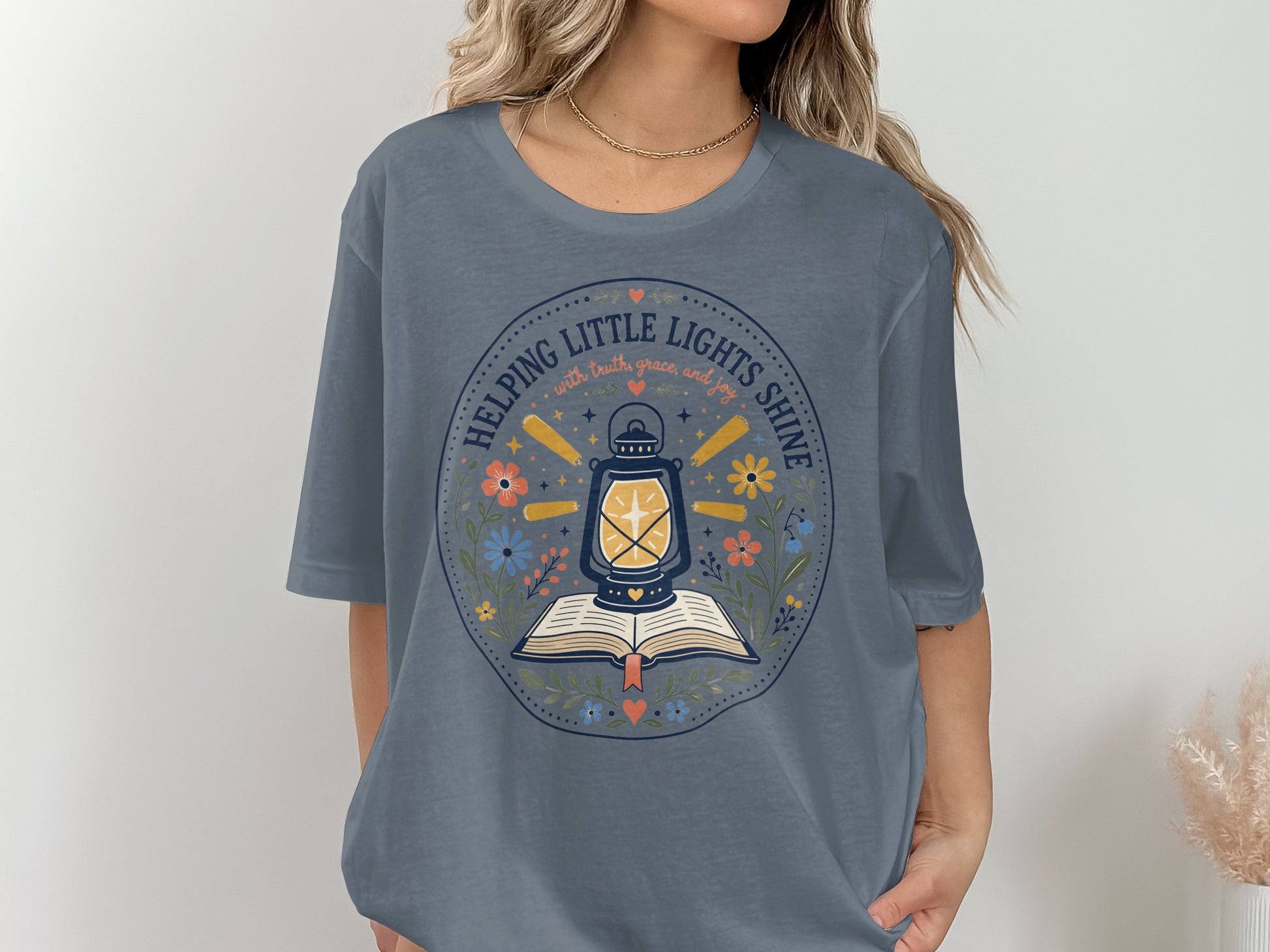 Blue t-shirt with a circular graphic of a lantern over an open book, surrounded by flowers and text.