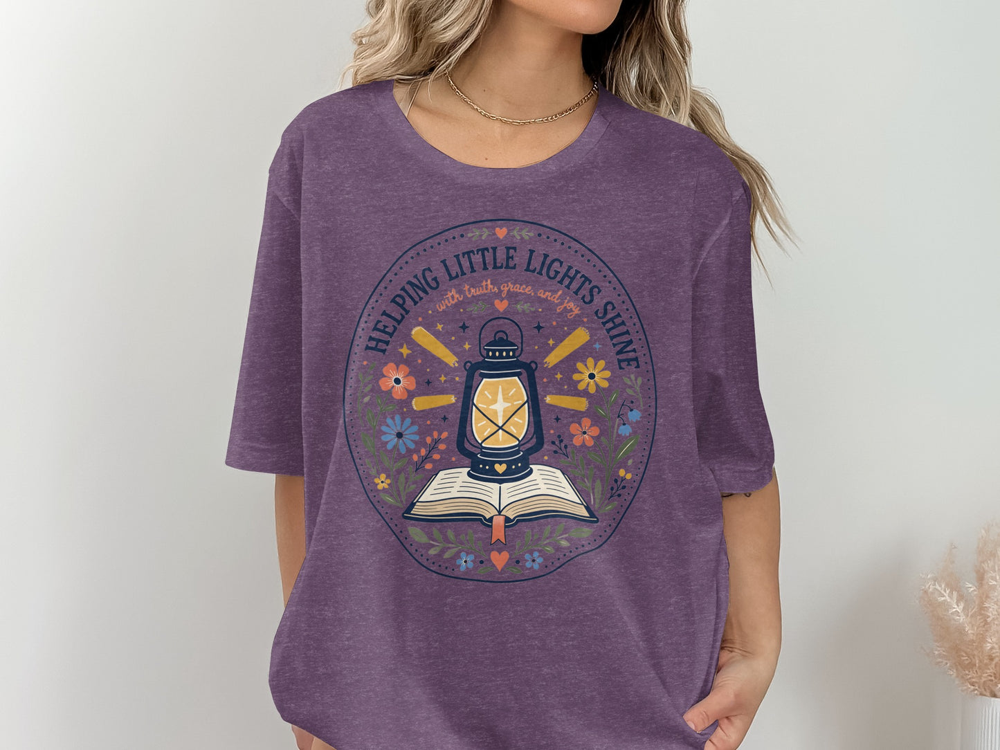 Purple t-shirt graphic with lantern on open book, surrounded by flowers and text.