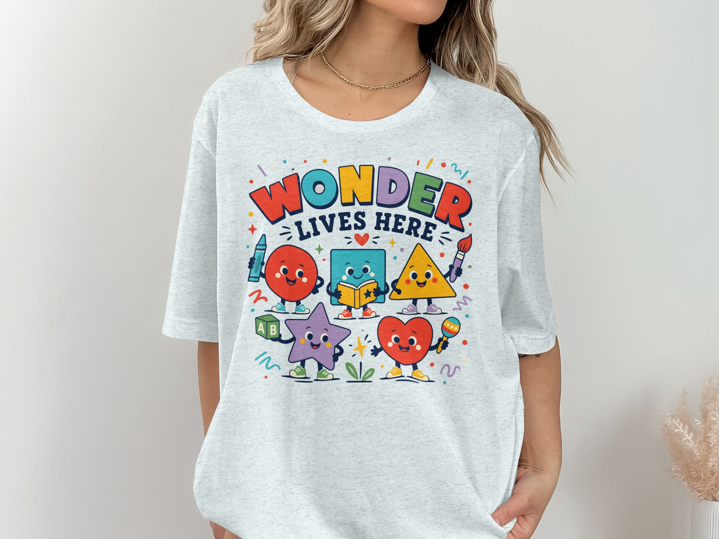 "Wonder Lives Here" text with cute cartoon shapes and school supplies on a light grey t-shirt.