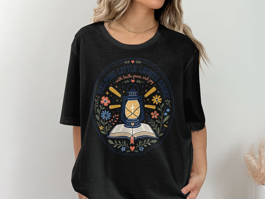 Black t-shirt with circular graphic of an open book, lantern, and flowers. Text says "Helping Little Lights Shine".