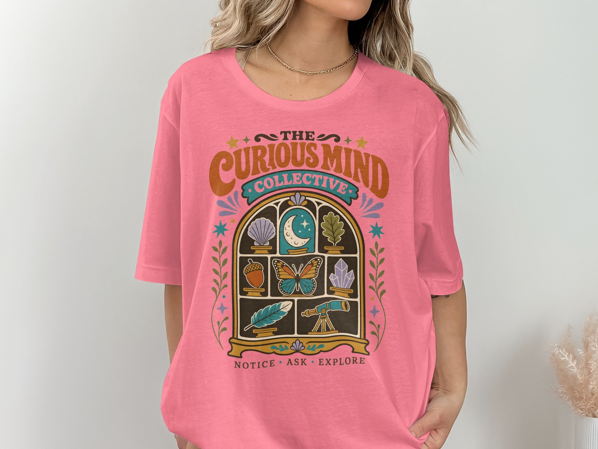 Pink t-shirt features "The Curious Mind Collective" logo with moon, butterfly, feather, and telescope.