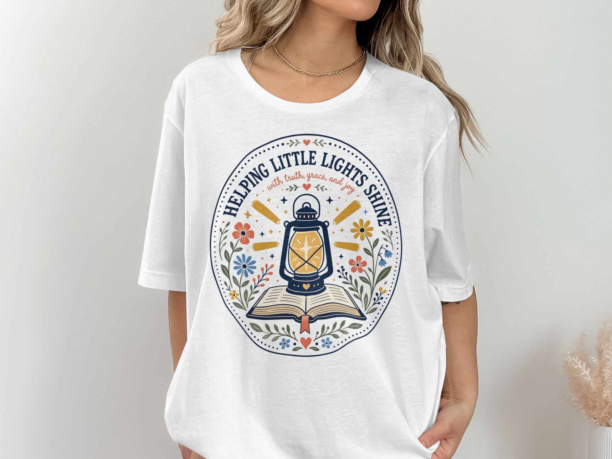 White t-shirt with graphic of lantern on open book, "Helping Little Lights Shine" text.