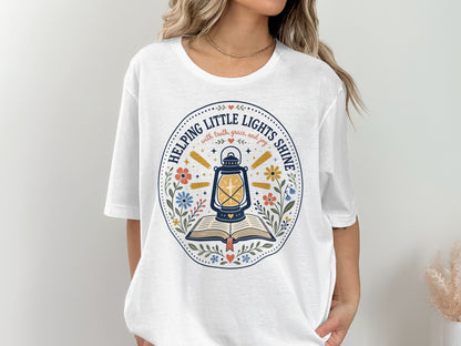 White t-shirt with graphic of lantern on open book, "Helping Little Lights Shine" text.