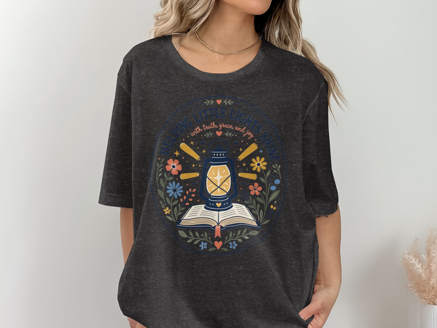 Dark grey t-shirt with circular graphic reading "HELPING LITTLE LIGHTS SHINE" and a lantern over a book.