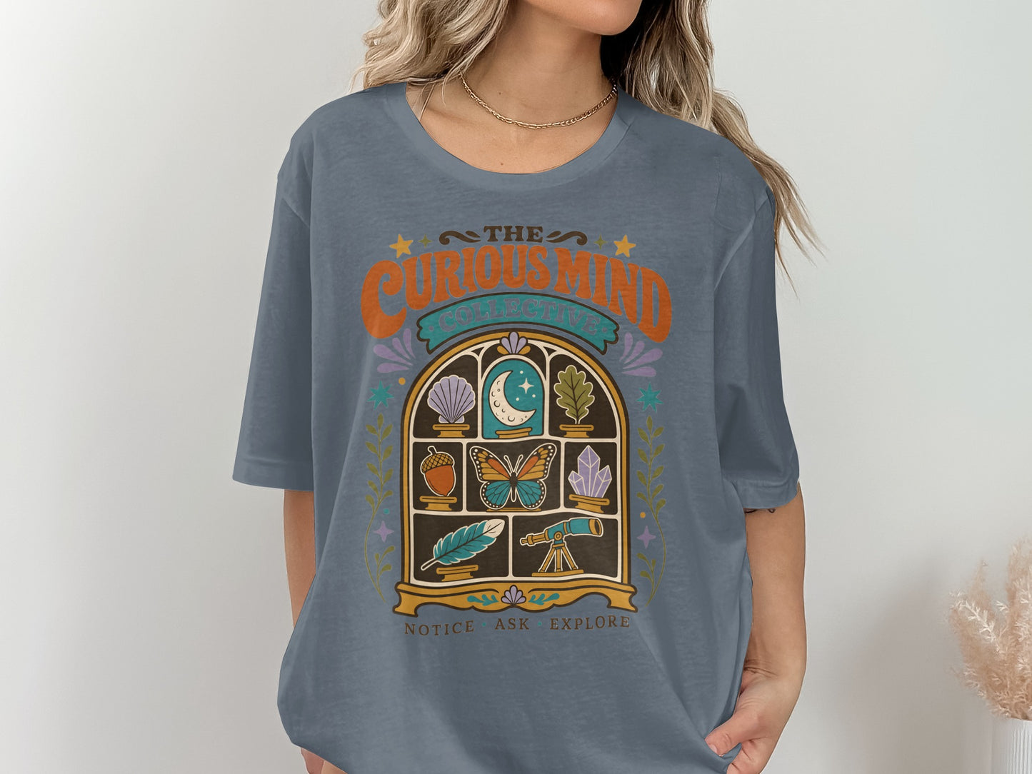 Blue t-shirt with "The Curious Mind Collective" text and nature-themed window graphic.