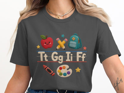 Graphic tee with apple, backpack, pencils, letters, crayon, and paint palette.