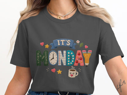"It's Monday" text with a clock, pencil, and cute designs on a dark gray t-shirt.