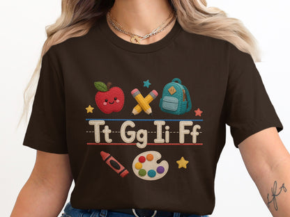 Brown t-shirt with embroidered cartoon apple, pencils, backpack, letters, crayon, and paint palette.