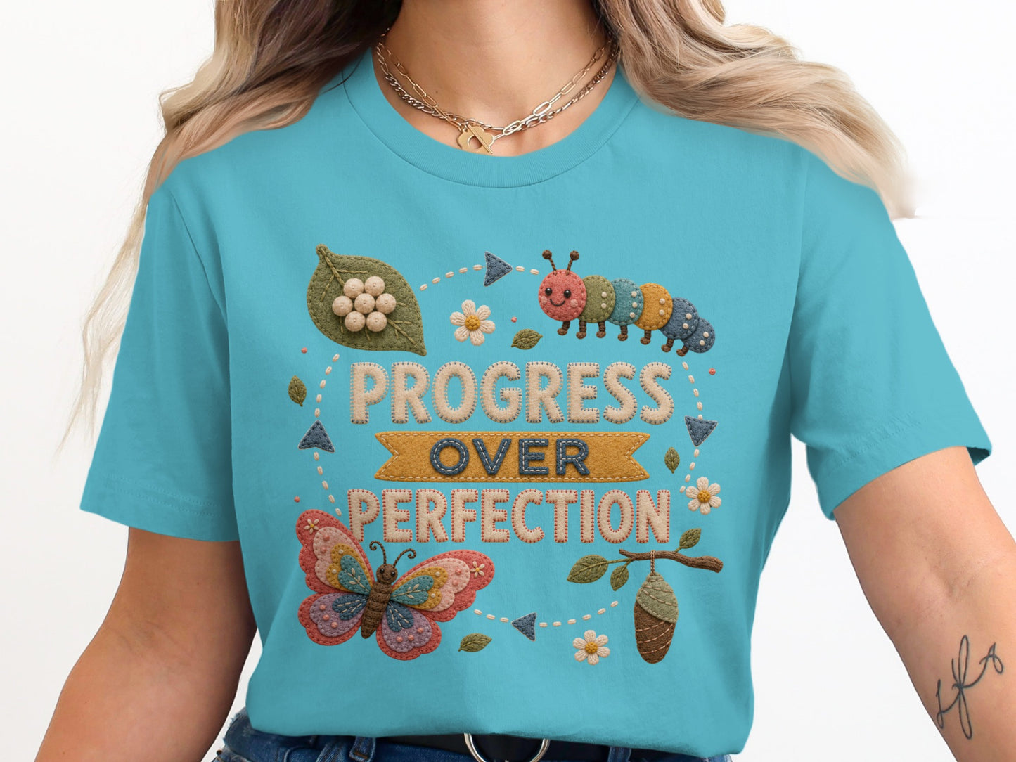 "Progress Over Perfection" text with butterfly, caterpillar, cocoon, and leaf motifs on a teal shirt.