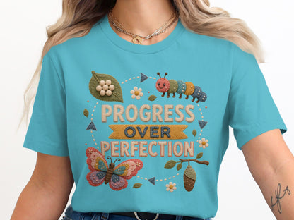 "Progress Over Perfection" text with butterfly, caterpillar, cocoon, and leaf motifs on a teal shirt.